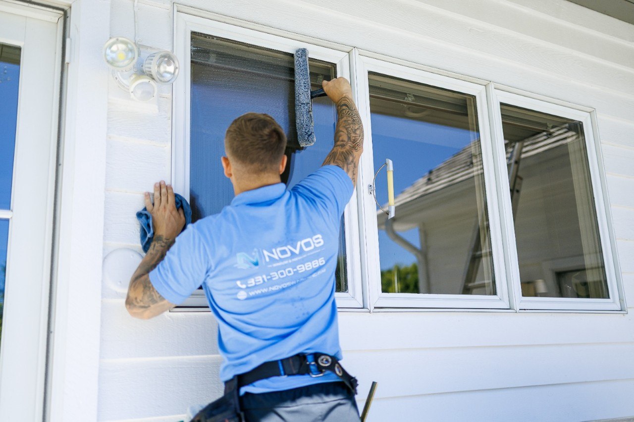 NOVOS technician hand-cleaning a three-pane window with a scrubber wand and microfiber cloth