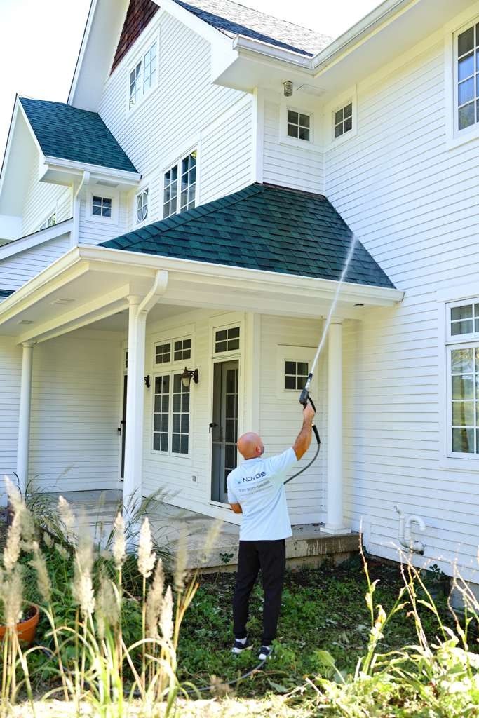 NOVOS technician in branded workwear pressure washing white lap siding above a porch roof