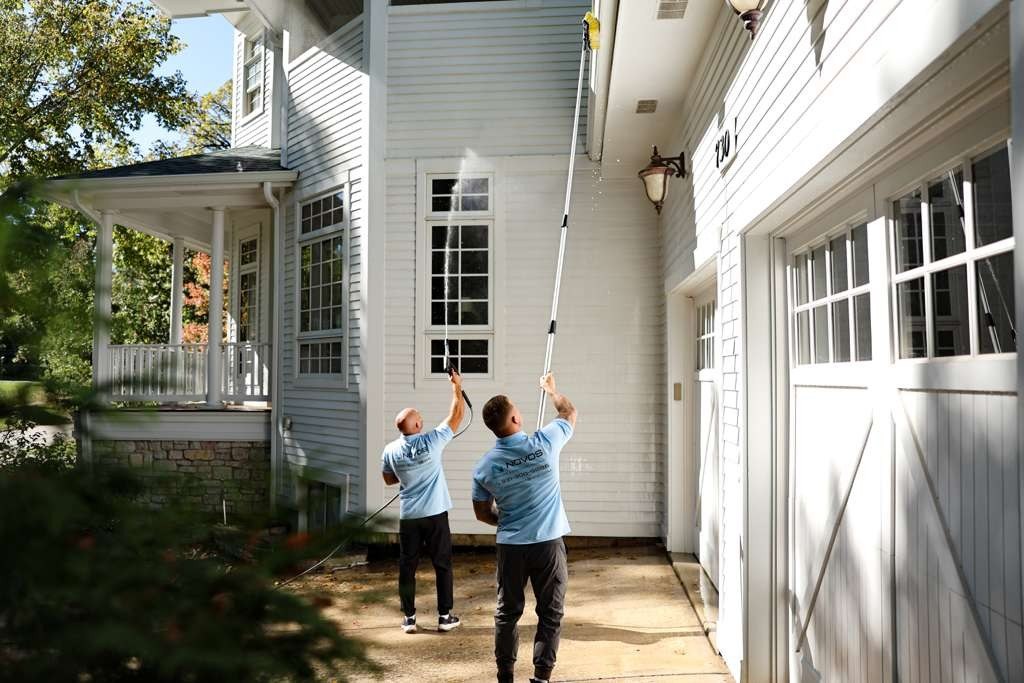 Two NOVOS technicians in branded workwear pressure washing and brushing the siding and roofline of a two-story home