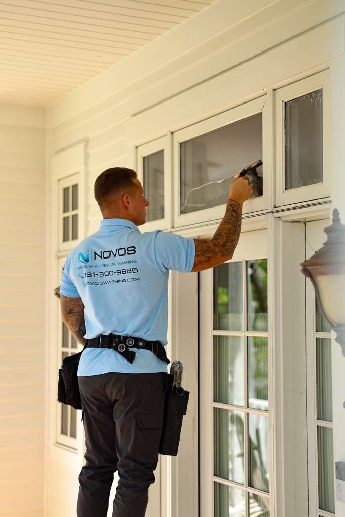 NOVOS technician in branded workwear squeegeeing a transom window above exterior glass doors